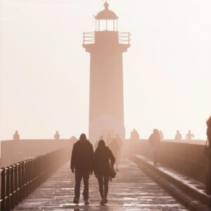 A romantic scene of a couple walking towards a lighthouse during sunset, with silhouettes and atmospheric lighting.